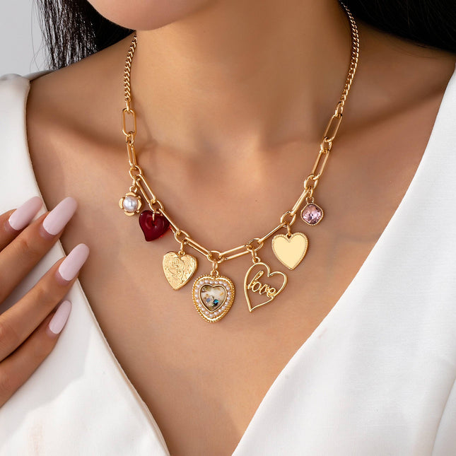 Gold plated elongated paperclip chain necklace with dangling porcelain heart, rhinestone crystal, pearl cluster, and red enamel heart charms styled in a romantic feminine jewelry aesthetic.
