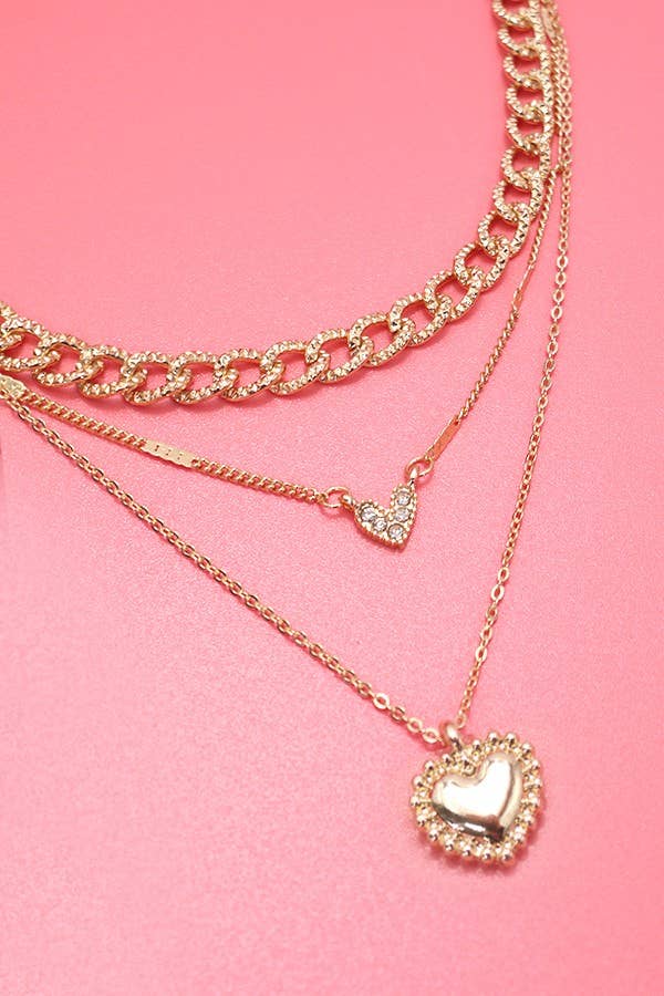 Gold layered necklace featuring multiple chains with rhinestone heart charms styled on a soft pink background.