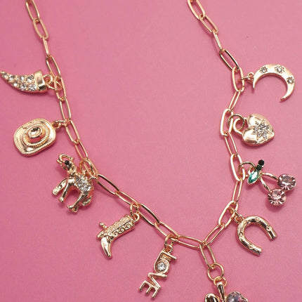 Gold western charm necklace featuring boots, cowboy hat, horseshoe, and heart charms styled on a pink background.