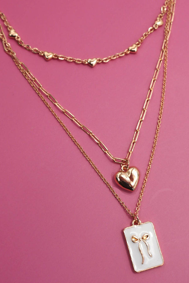 Layered gold necklaces with heart and rectangular pendants on a pink background