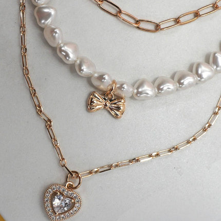 Gold layered necklace featuring pearl strand, bow charm, and rhinestone heart pendant styled on a neutral background