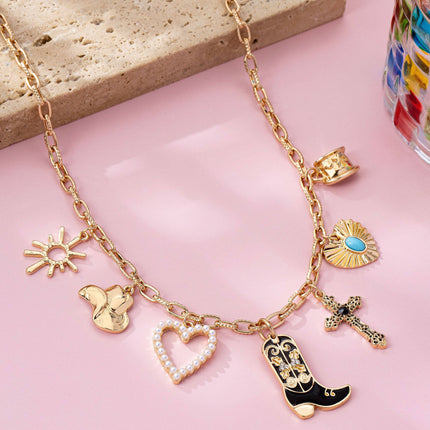 Gold paperclip chain necklace with western-inspired charms including a cowboy boot, heart, cross, sunburst, and hat, styled on a model’s neckline against a soft neutral background