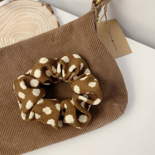 Brown scrunchie with white polka dots on a brown textured bag with a visible brand tag.