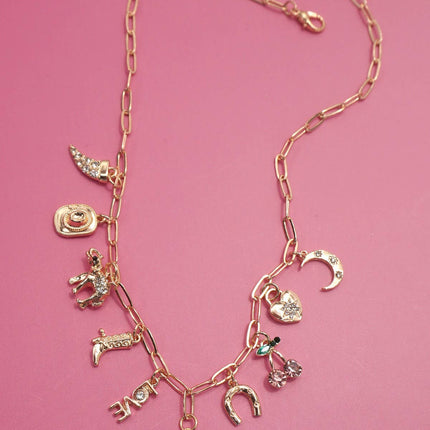Gold western charm necklace featuring boots, cowboy hat, horseshoe, and heart charms styled on a pink background.