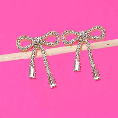 Gold rhinestone bow drop earrings with a delicate dangling design, shown worn on the ear and displayed against a bright pink background.