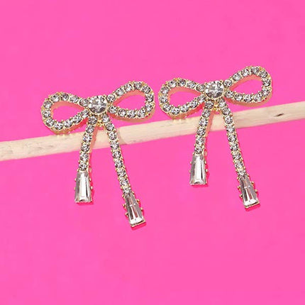 Gold rhinestone bow drop earrings with a delicate dangling design, shown worn on the ear and displayed against a bright pink background.
