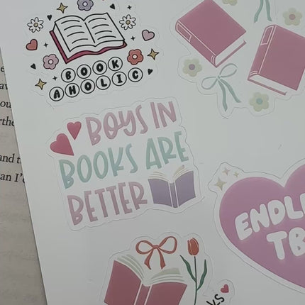 Multiple book lover sticker sheet designs featuring cozy reading themes, spicy book humor, hearts, books, and playful illustrations displayed on an open book background.