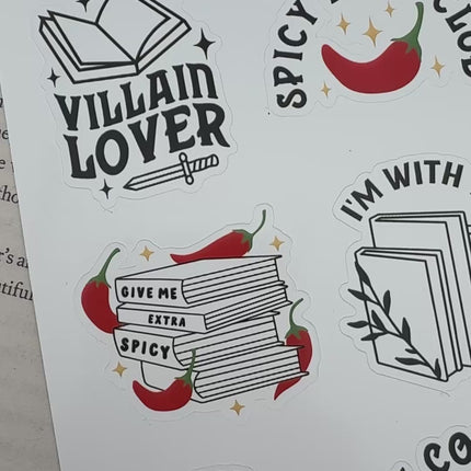 Multiple book lover sticker sheet designs featuring cozy reading themes, spicy book humor, hearts, books, and playful illustrations displayed on an open book background.