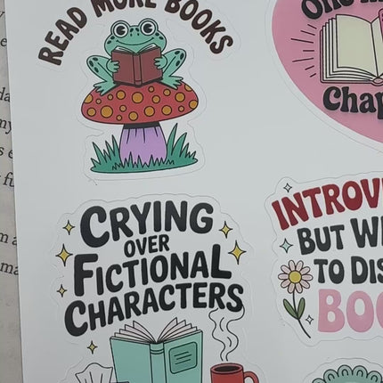 Multiple book lover sticker sheet designs featuring cozy reading themes, spicy book humor, hearts, books, and playful illustrations displayed on an open book background.