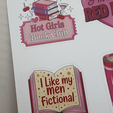 Multiple book lover sticker sheet designs featuring cozy reading themes, spicy book humor, hearts, books, and playful illustrations displayed on an open book background.