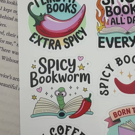 Multiple book lover sticker sheet designs featuring cozy reading themes, spicy book humor, hearts, books, and playful illustrations displayed on an open book background.