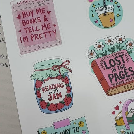 Multiple book lover sticker sheet designs featuring cozy reading themes, spicy book humor, hearts, books, and playful illustrations displayed on an open book background.