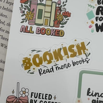 Multiple book lover sticker sheet designs featuring cozy reading themes, spicy book humor, hearts, books, and playful illustrations displayed on an open book background.